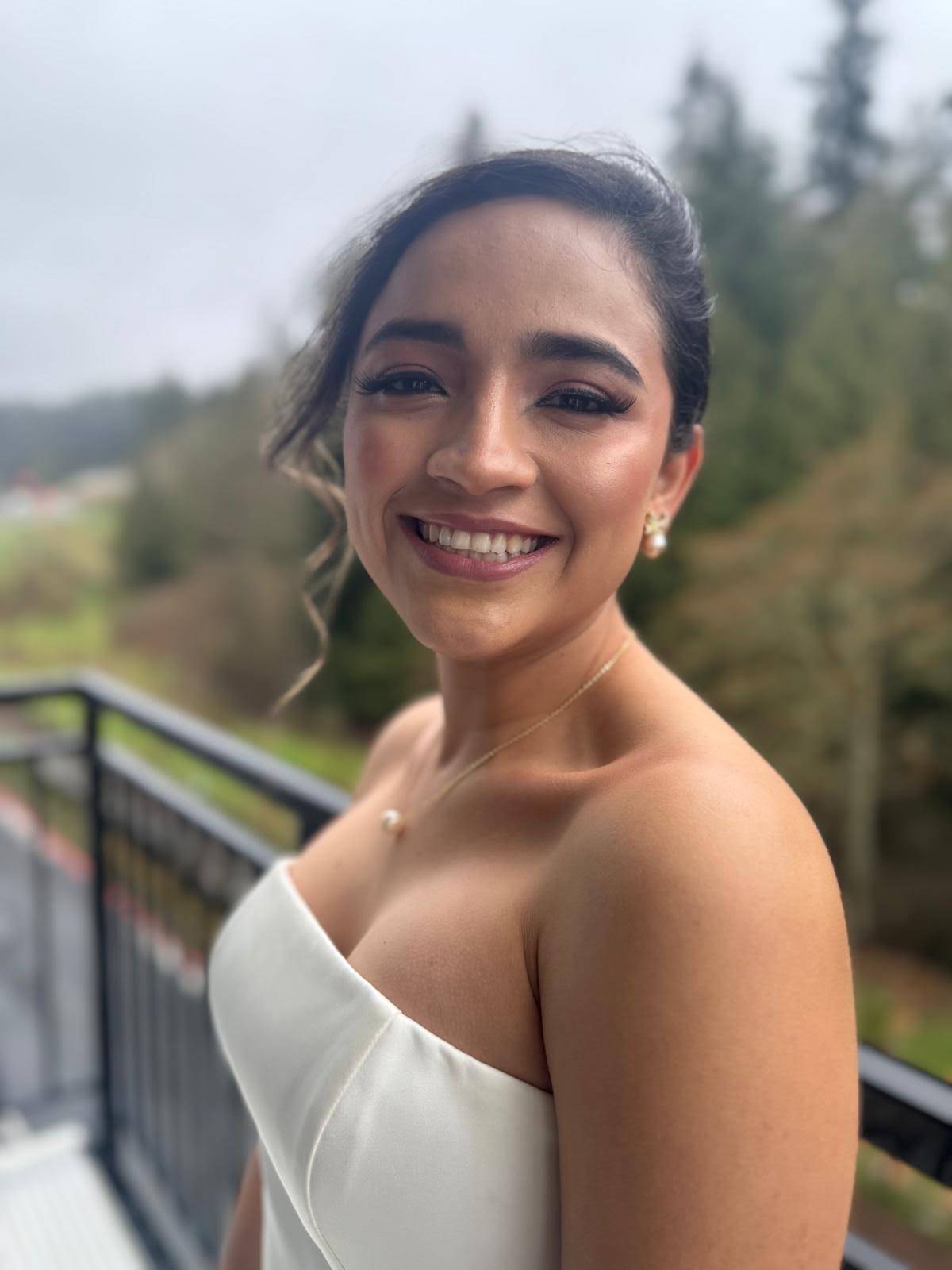 Bride in a strapless ivory gown smiling outdoors against Pacific Northwest evergreens, soft glam by Eleglam